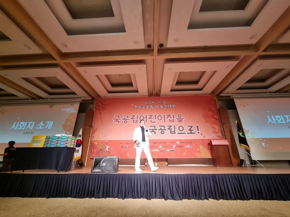 연회장 무대에서 흰 옷을 입은 MC가 진행하고 뒤에 대회 백드롭과 스크린이 있는 모습