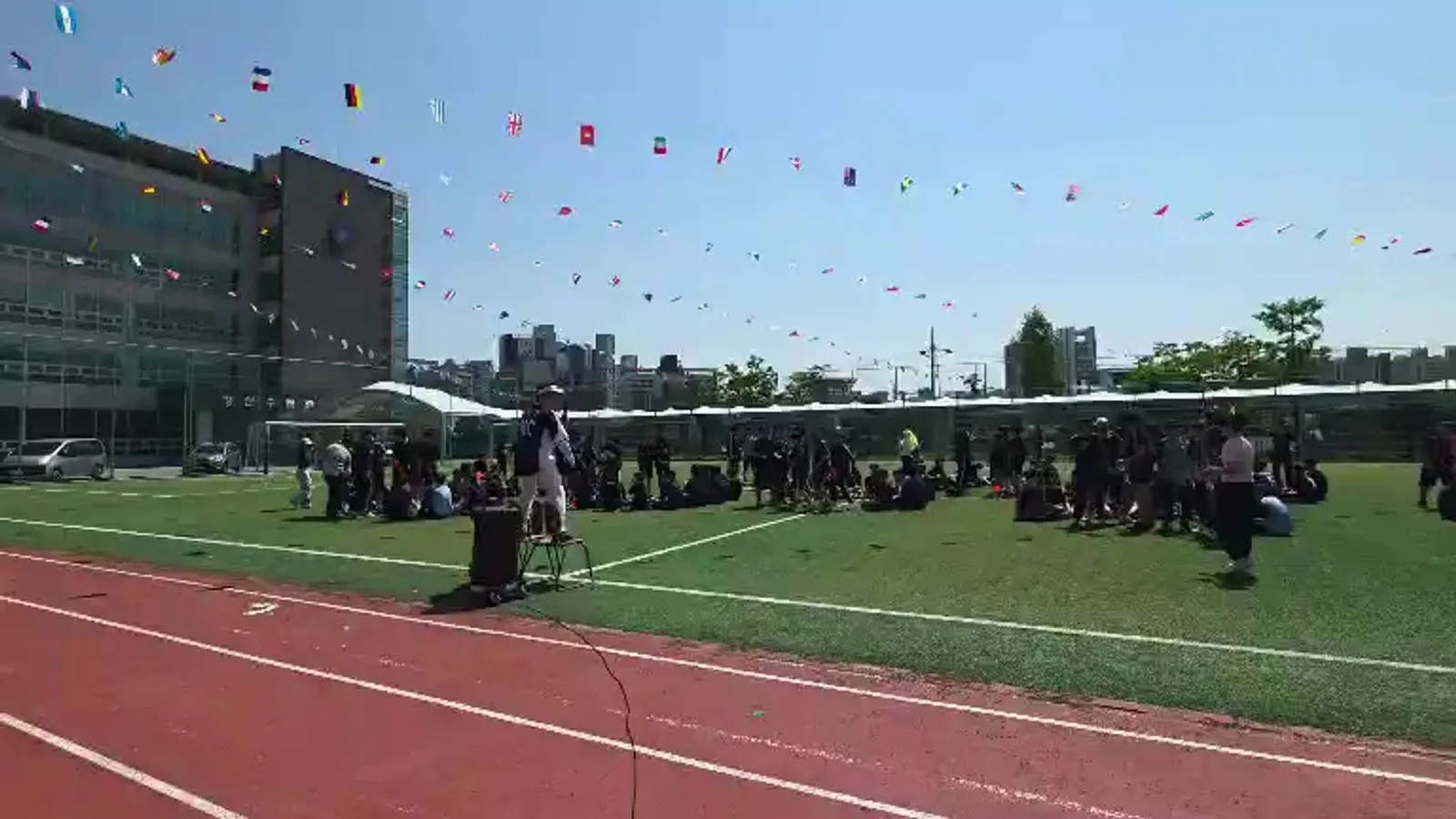 운동장 트랙 옆에서 진행자가 마이크를 잡고 학생들이 모인 모습을 멀리서 찍은 사진