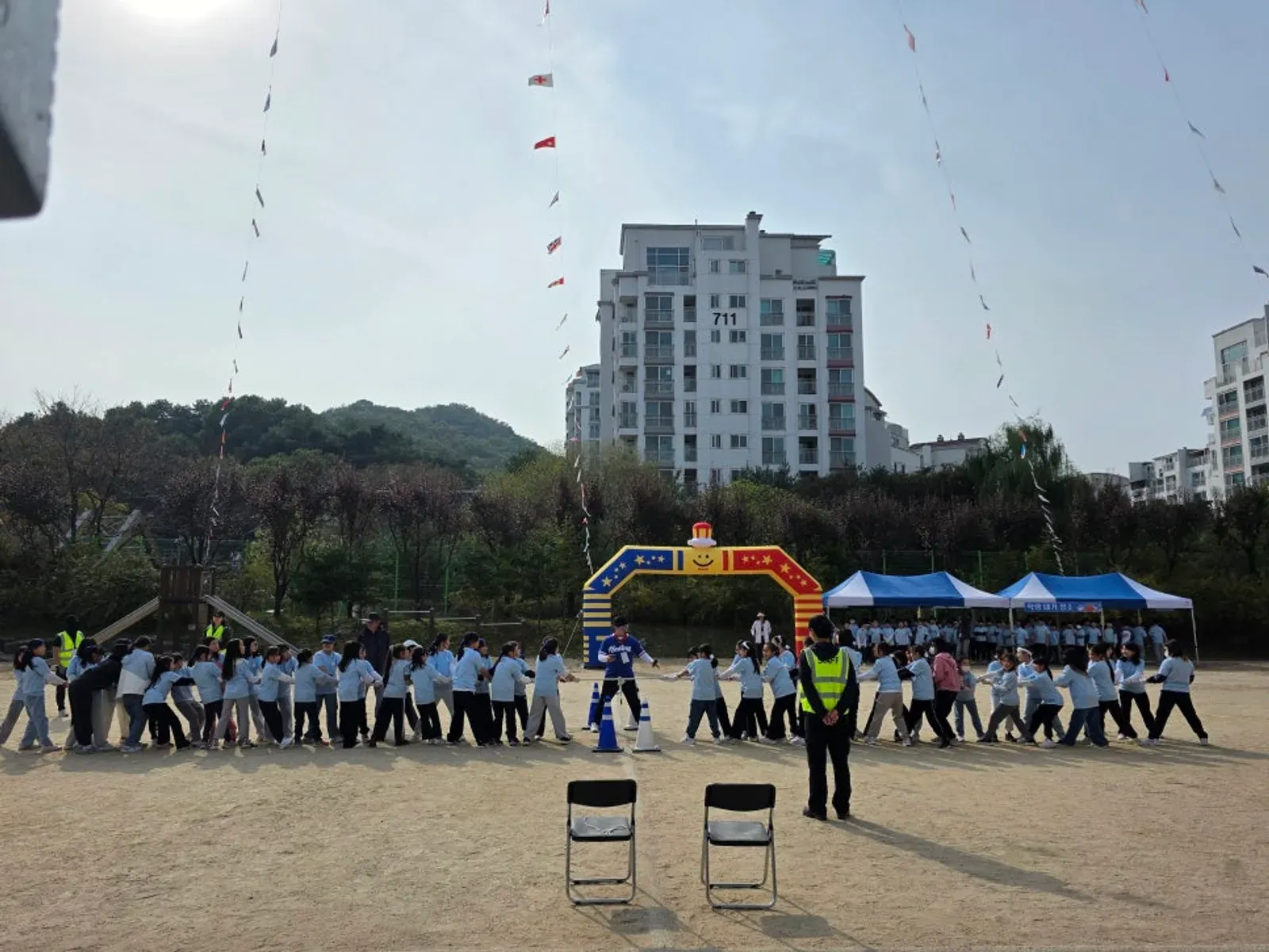 풍선 아치 앞에서 학생들이 어깨를 잡고 줄지어 걷고 안전조끼 입은 요원이 지켜보는 모습