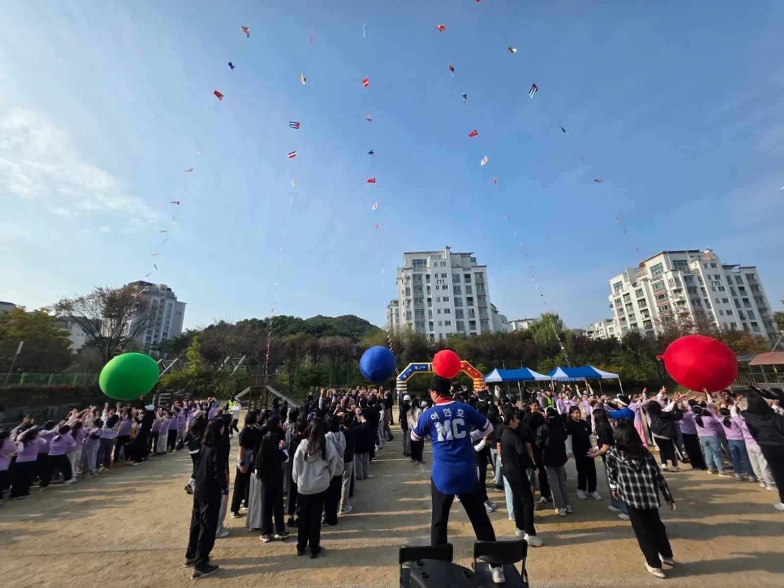 MC 조끼를 입은 진행자가 운동장 가운데서 학생들과 함께 팔을 드는 모습