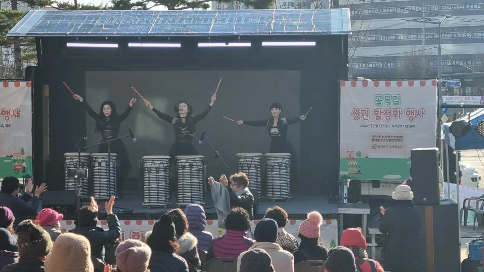 거리 축제 무대에서 북 공연이 열리고 앞쪽에 털모자를 쓴 관객들이 앉아 있는 모습